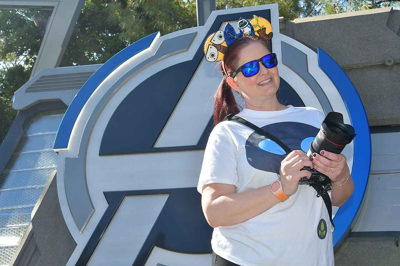 Someone wearing a white t-shirt, MICKEY MOUSE EARS and sunglasses poses with a camera in front of a blue circular wall display.