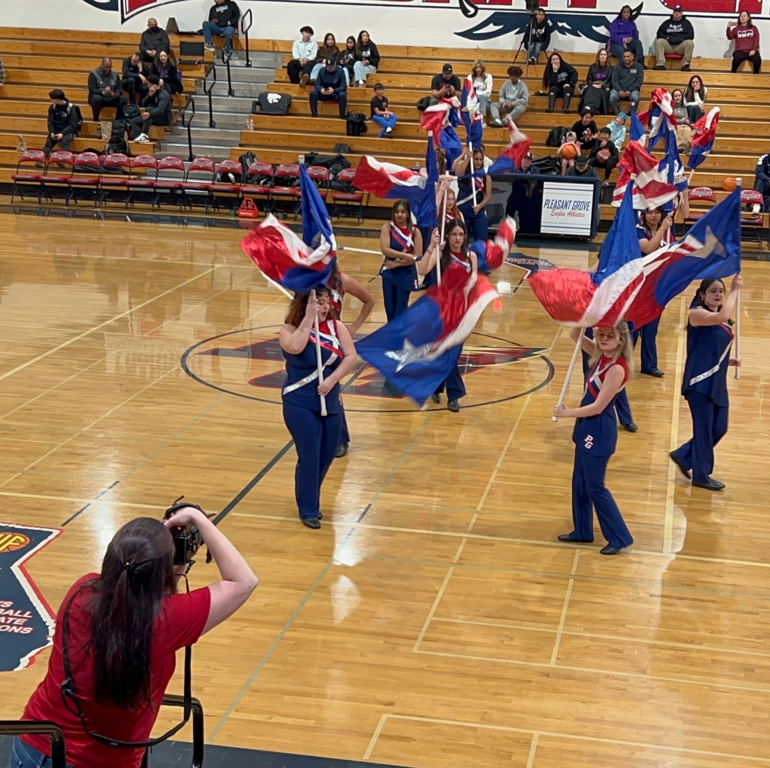 Color guard performers in blue uniforms practice their routine with flags on a gymnasium floor during rehearsal.