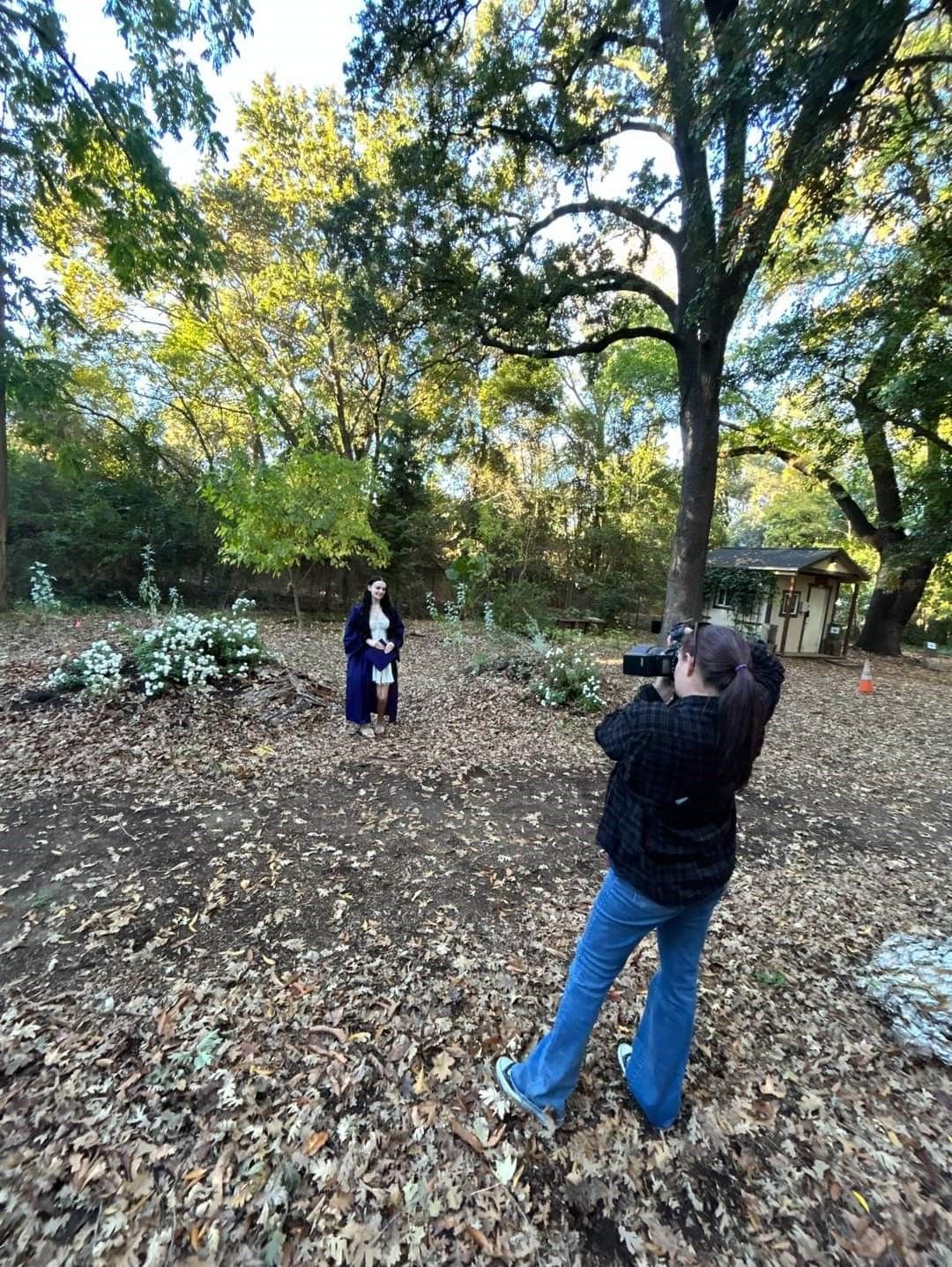 A photographer takes pictures in a leaf-covered outdoor area with trees and natural lighting.