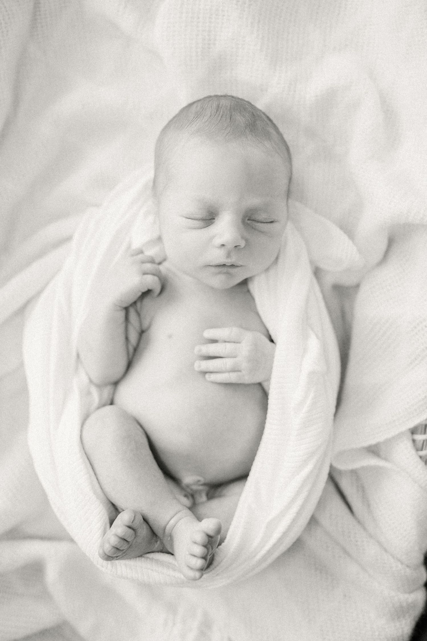 Black and white portrait of a sleeping newborn baby wrapped in soft white fabric against light bedding.