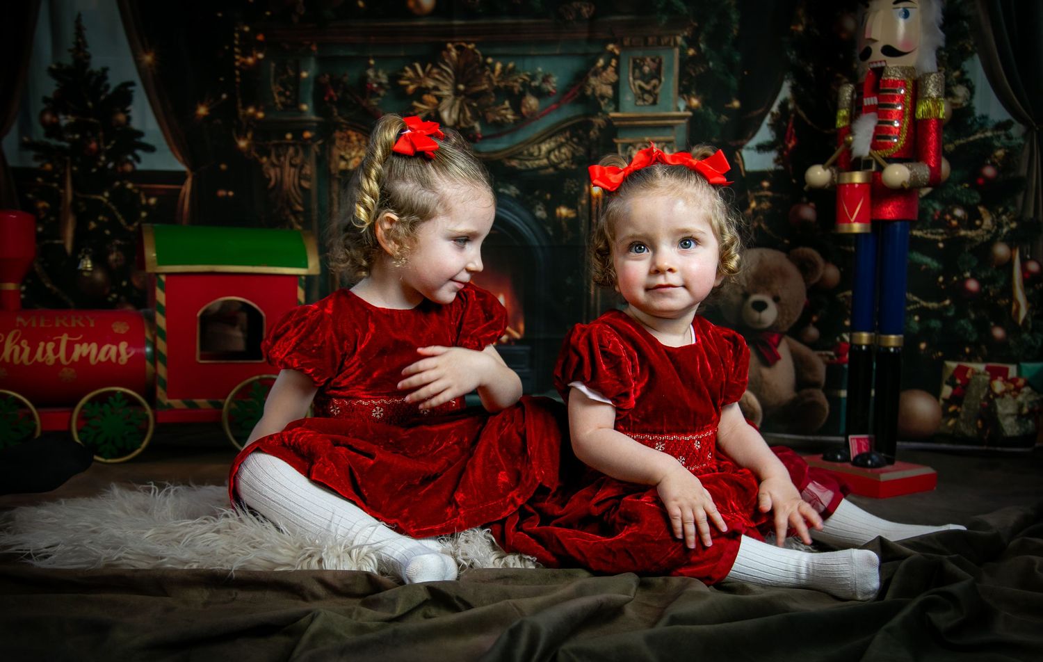 Two children in red velvet dresses sit on floor by Christmas tree with toy train and nutcracker decorations.
