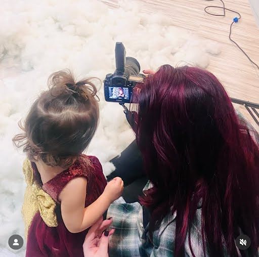 A person with burgundy hair films content with a small child on a wooden floor using a digital camera.