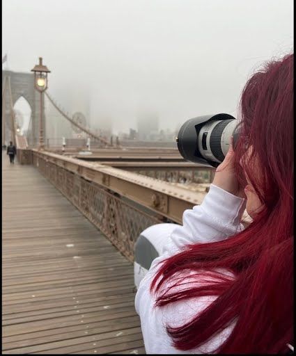 Photographer with bright red hair takes pictures on a foggy bridge walkway with vintage lampposts.