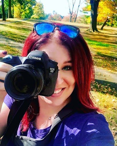 Photographer holds Canon DSLR camera outdoors in autumn park with sunglasses on head and purple shirt.