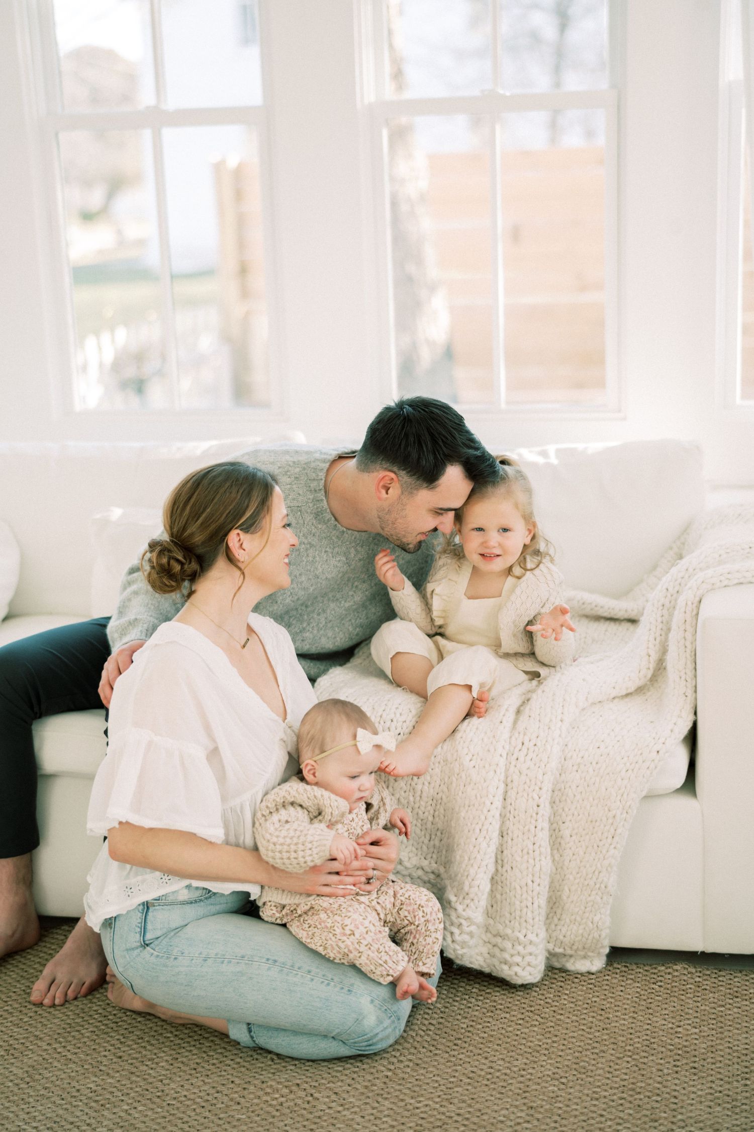 Family sharing tender moment together on white couch in bright sunlit room with neutral decor.