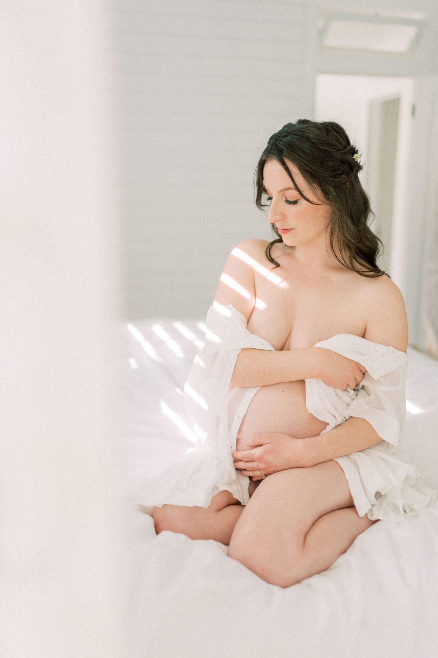A dreamy maternity photo shows an expectant mother in white sitting by sunlit windows.