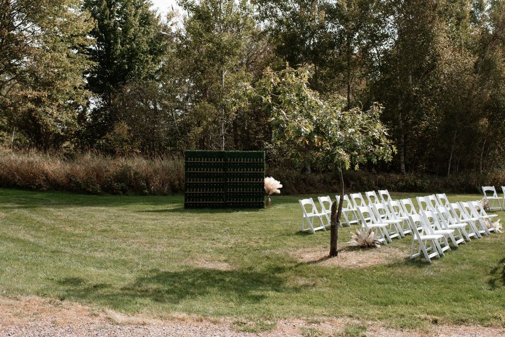 Outdoor wedding ceremony setup with white folding chairs and a black backdrop on a grassy lawn near trees.