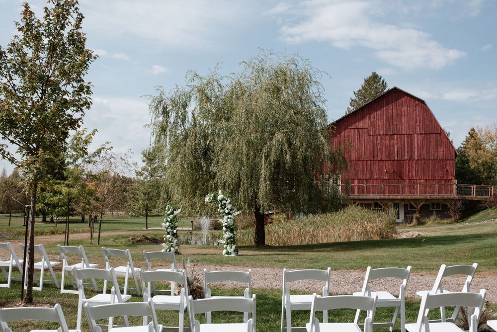 A rustic red barn and willow tree overlook white wedding chairs set up for an outdoor ceremony on a sunny day.
