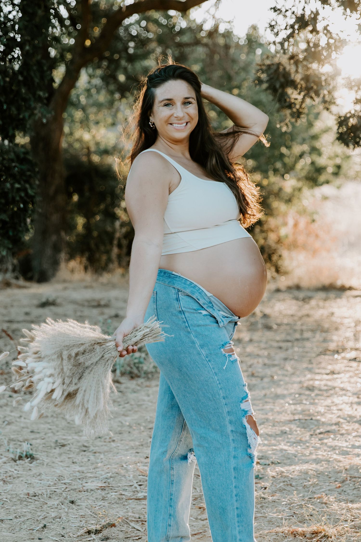 Expecting mother in white top and jeans poses with dried pampas grass in golden sunlight.