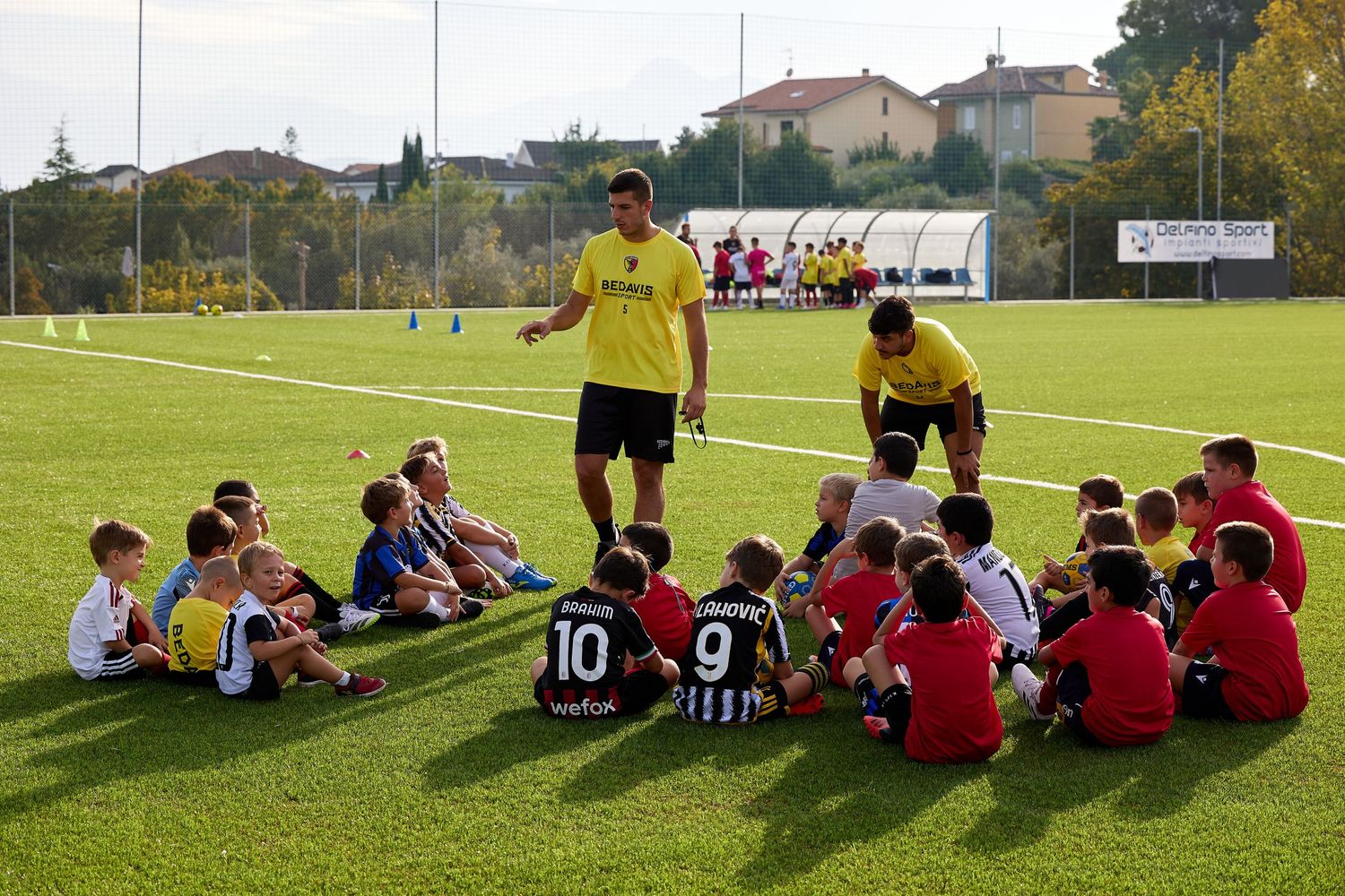 Youth soccer team sits on grass field listening to coaches during training session.