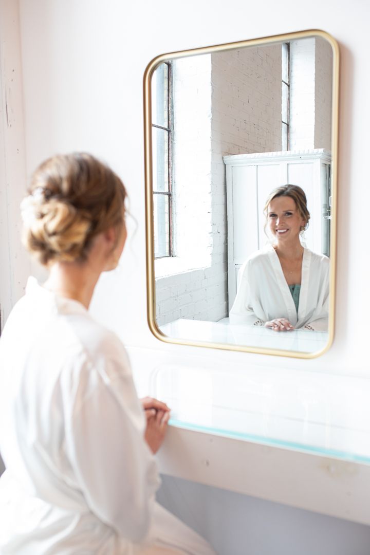 Bridal preparation reflected in modern gold-framed mirror against white brick wall.