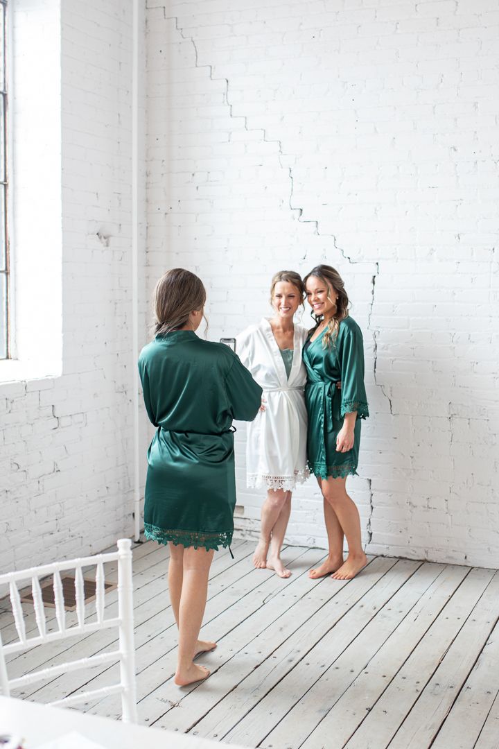 Three people in robes standing in a bright white room with wooden floors during wedding preparations.