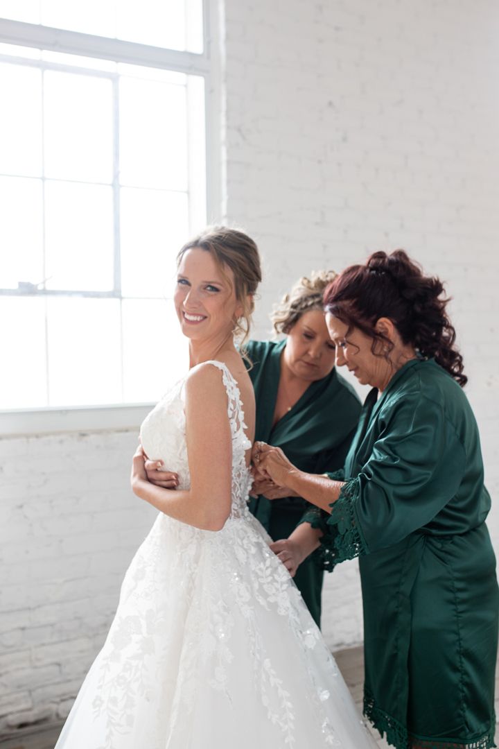 Final adjustments being made to a white lace wedding gown in a bright sunlit room.