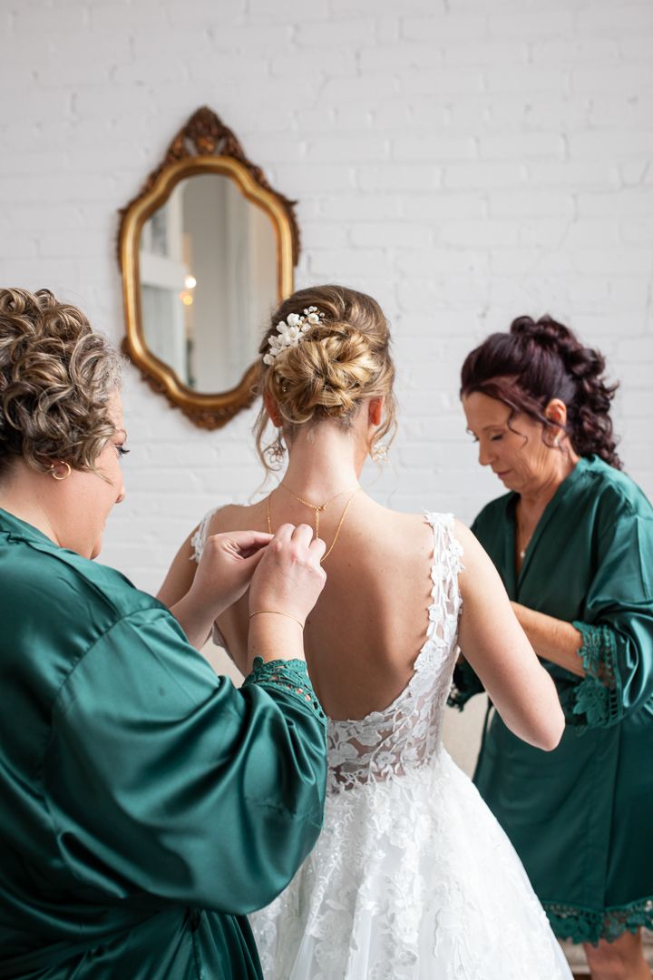 Assistants in emerald robes help secure the back of a detailed lace wedding dress.