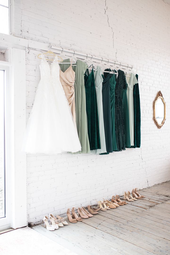 Bridal party dresses in white and green tones hanging on wall with shoes below.