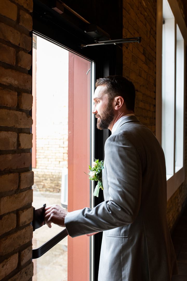 A suited figure gazes contemplatively through a window in a rustic brick interior.