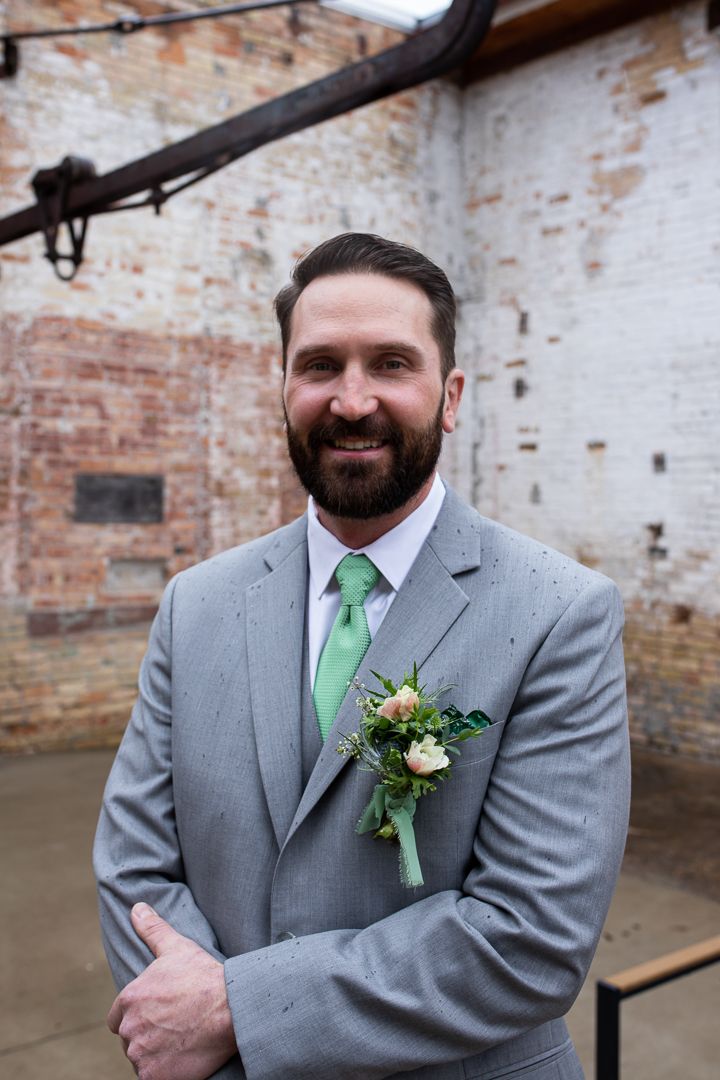 Groom in grey suit with green tie and boutonniere against brick wall.