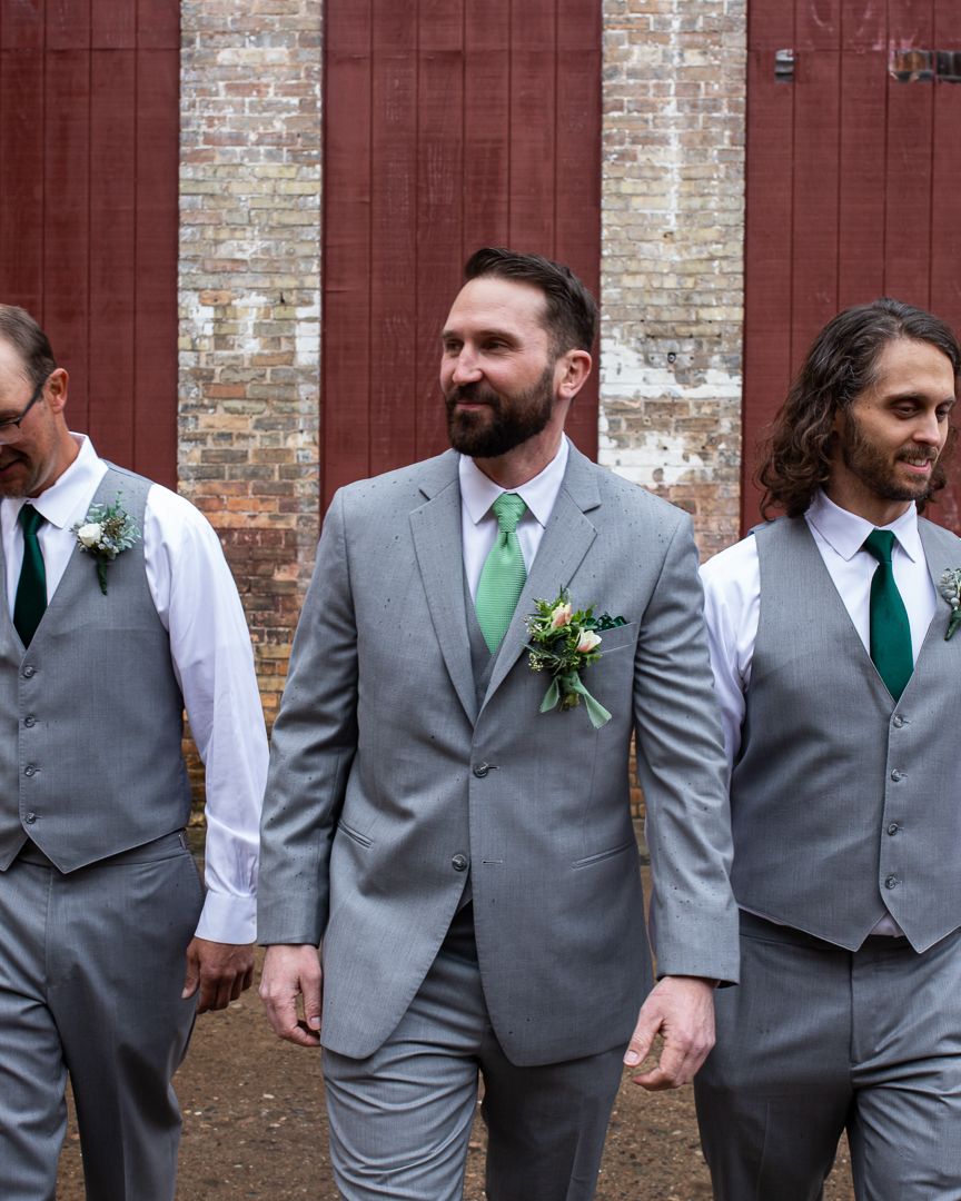 Groomsmen in coordinated gray suits and green ties pose together against a brick wall backdrop.