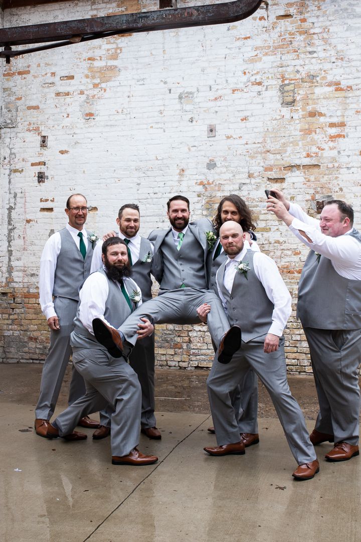 Wedding party in gray suits and green ties poses playfully against white brick wall.