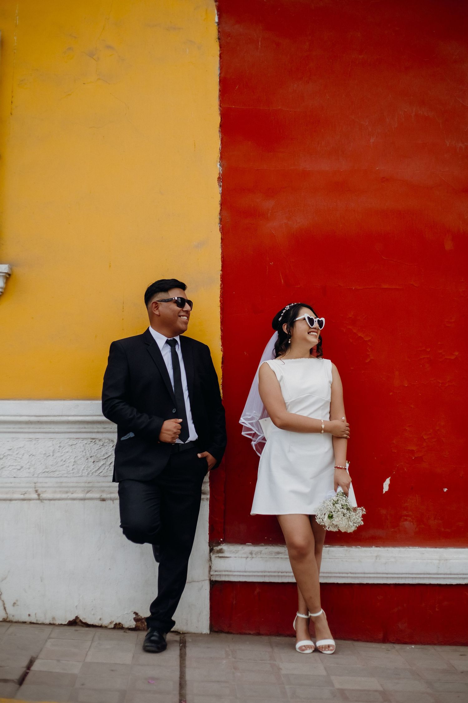 Wedding couple posing against vibrant yellow and red wall in urban setting.