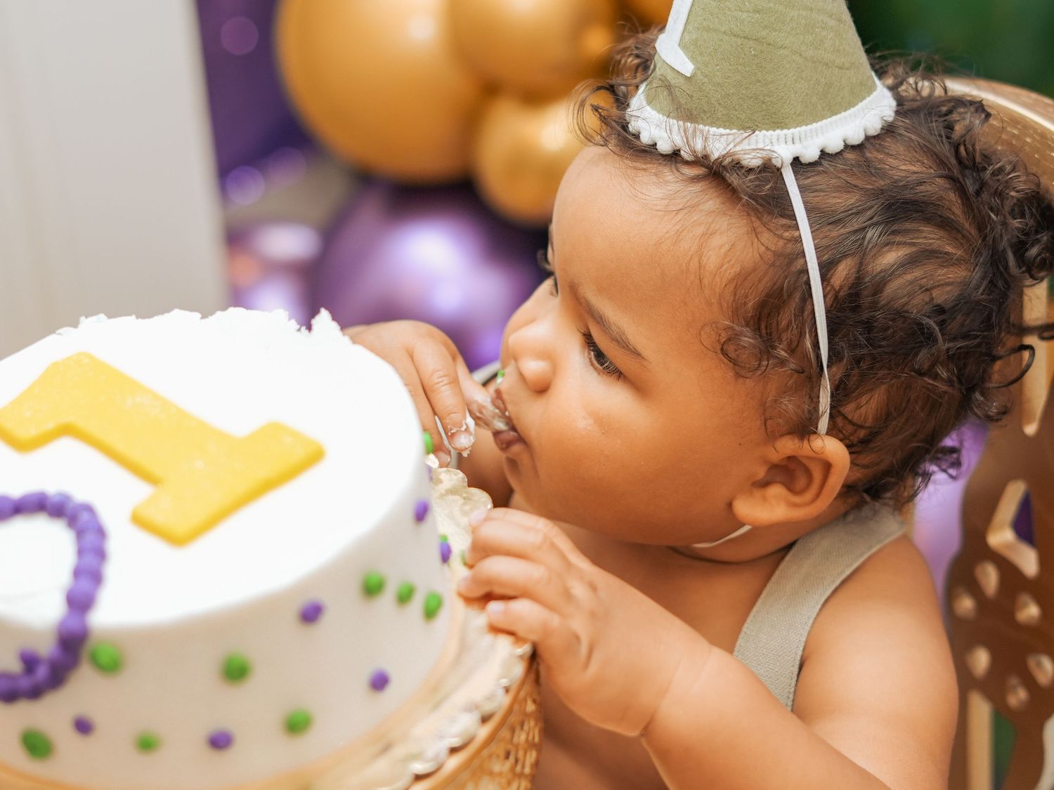 Young child blowing out a candle on a first birthday cake with purple and white decorations.