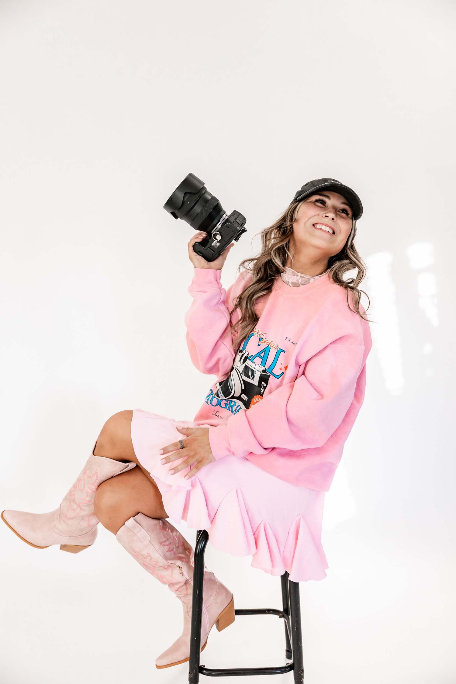 Wyoming Photographer
Professional photographer wearing pink sweater dress and boots sits on stool with camera in studio setting.