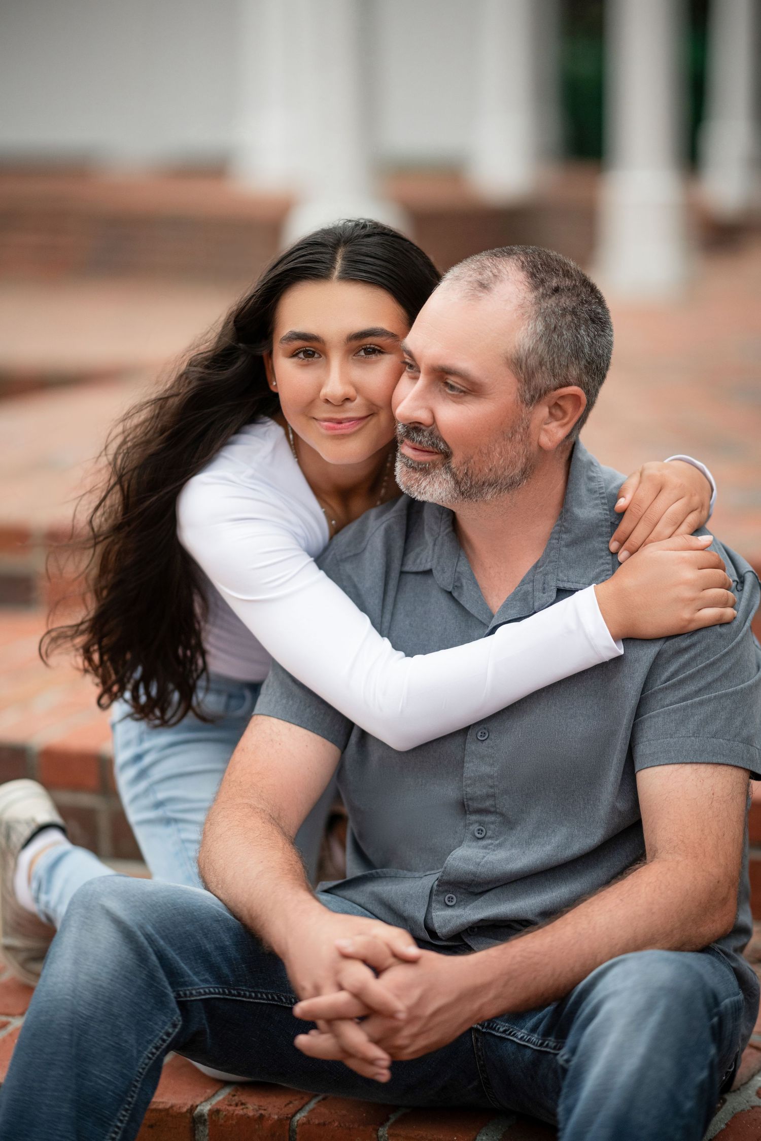 Two people embrace and smile while sitting on brick steps outdoors.