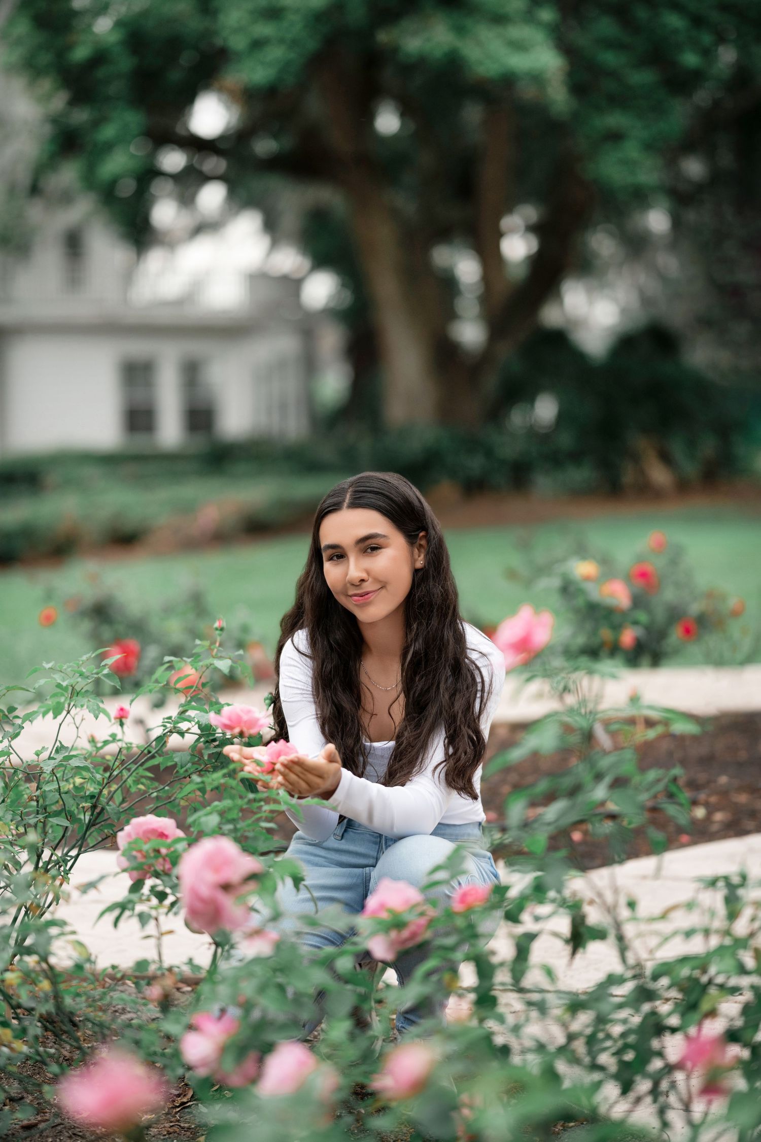 A person wearing a white top and jeans kneels in a blooming garden of pink roses on a summer day.