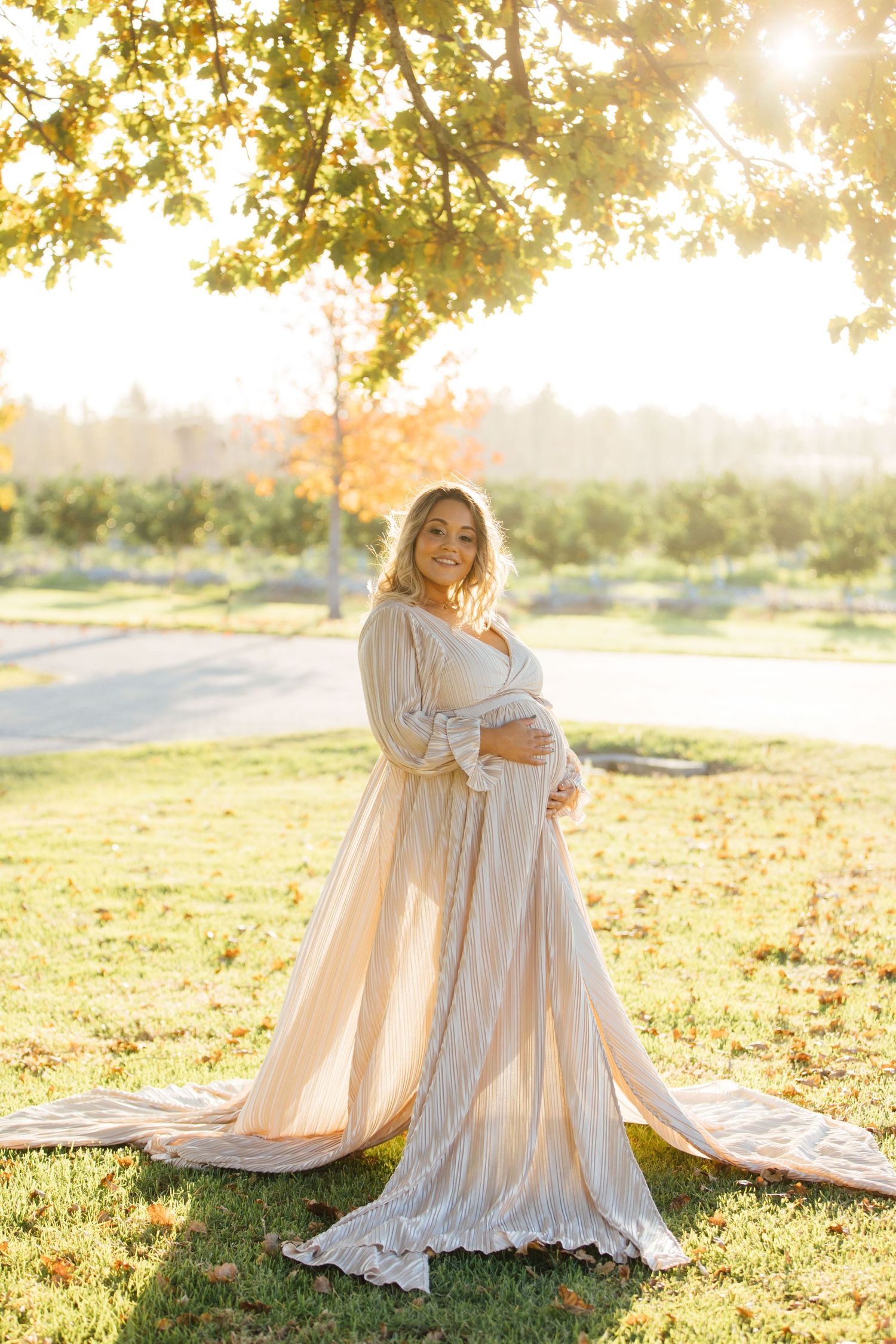 A flowing cream-colored dress catches golden sunlight in an outdoor maternity photoshoot amid autumn trees and grass.