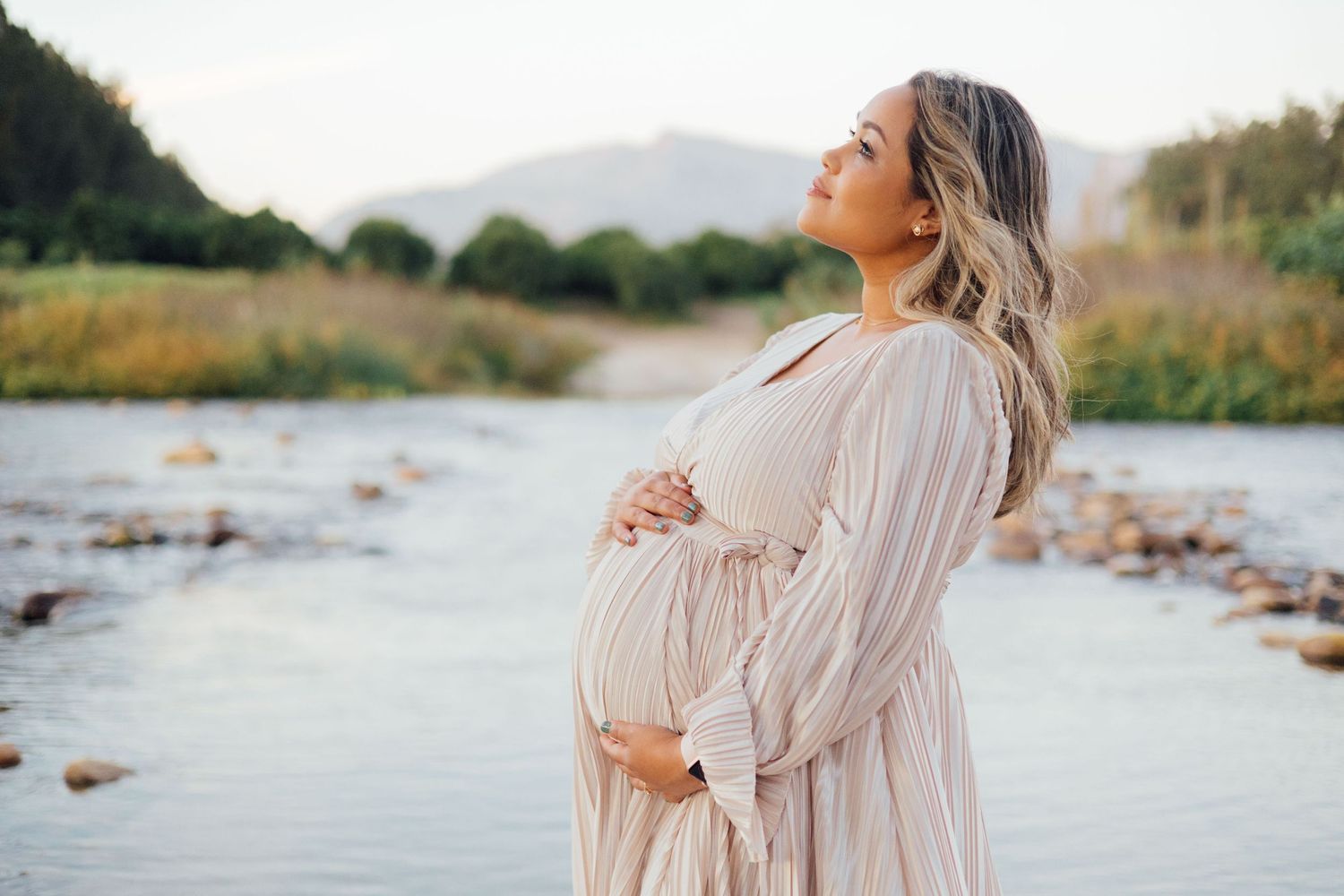 A pregnant woman in a flowy beige dress stands peacefully by a river with mountains in the background at sunset.