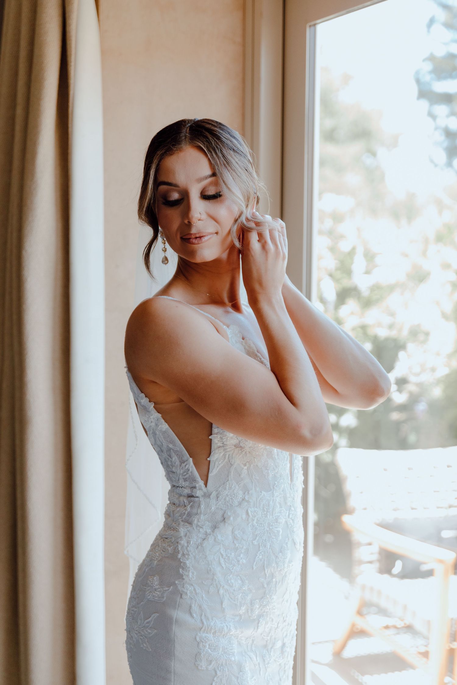 Bride wearing a fitted lace wedding dress standing by a sunlit window.