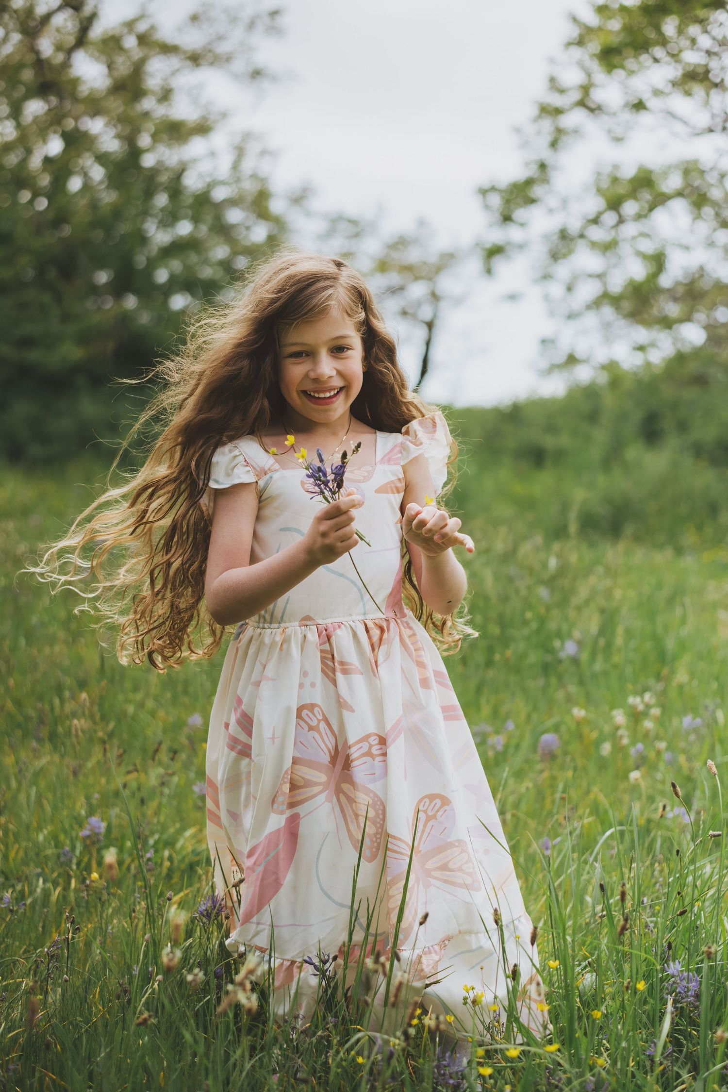 Young girl wearing a floral dress stands in a summer meadow with wildflowers while wind blows through her long hair.