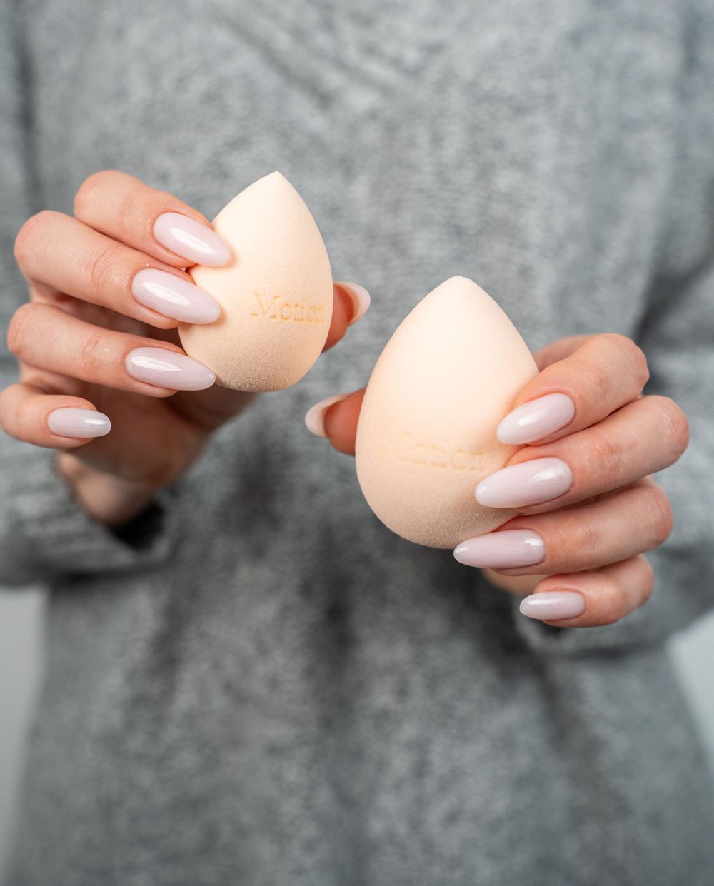 Manicured hands holding two peach-colored makeup blending sponges against a grey background.