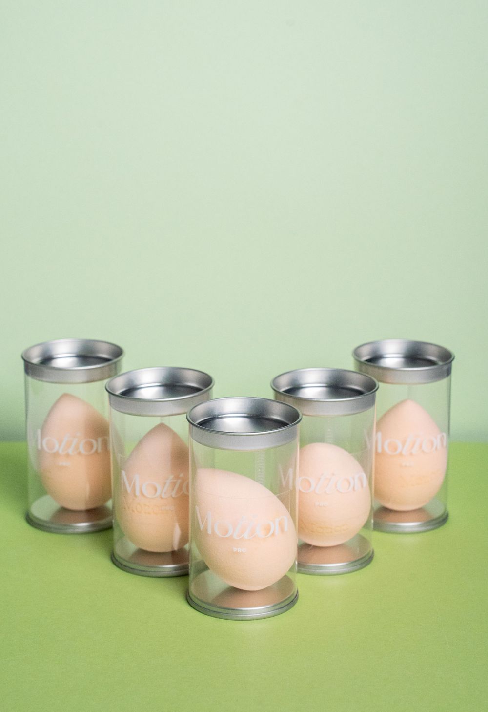 Five Motion makeup sponges in clear containers arranged in a curved formation on green surface.