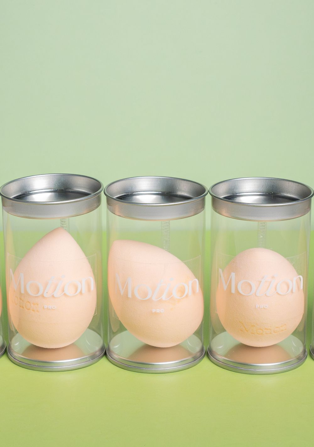 Three Motion makeup sponges displayed in clear cylindrical containers against lime green background.