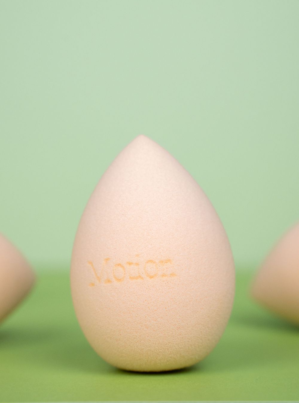 Peach-colored teardrop shaped makeup sponge with Motion logo embossed against mint green background.