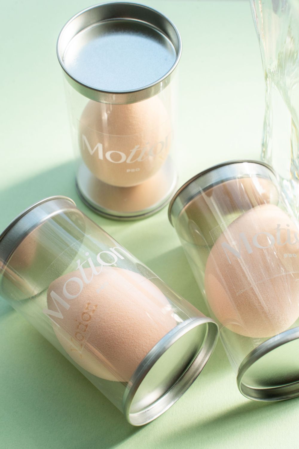 Three clear cylindrical containers with peach Motion makeup sponges arranged on mint green surface.