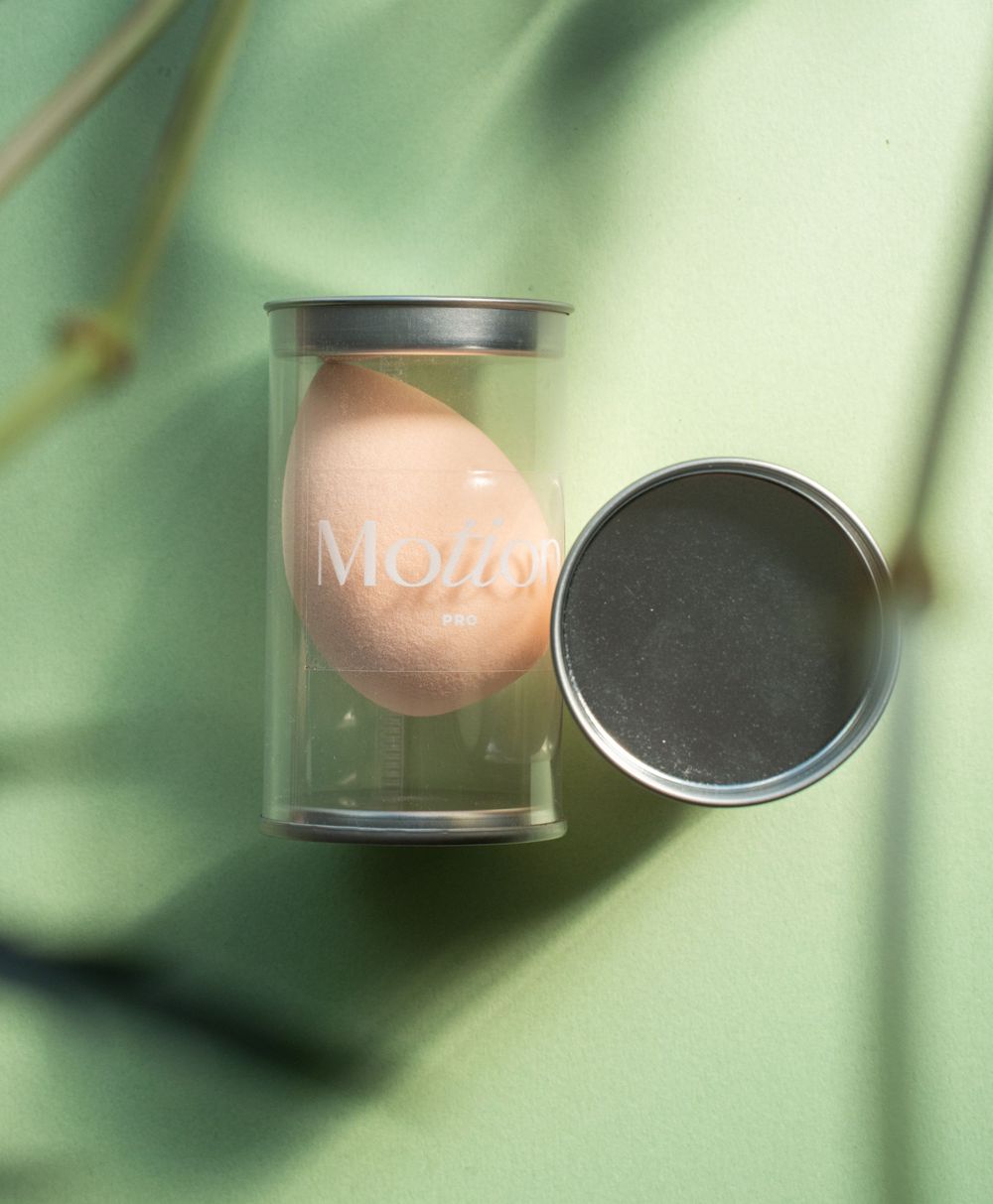 Motion makeup sponge in clear container with metal lid against green background.