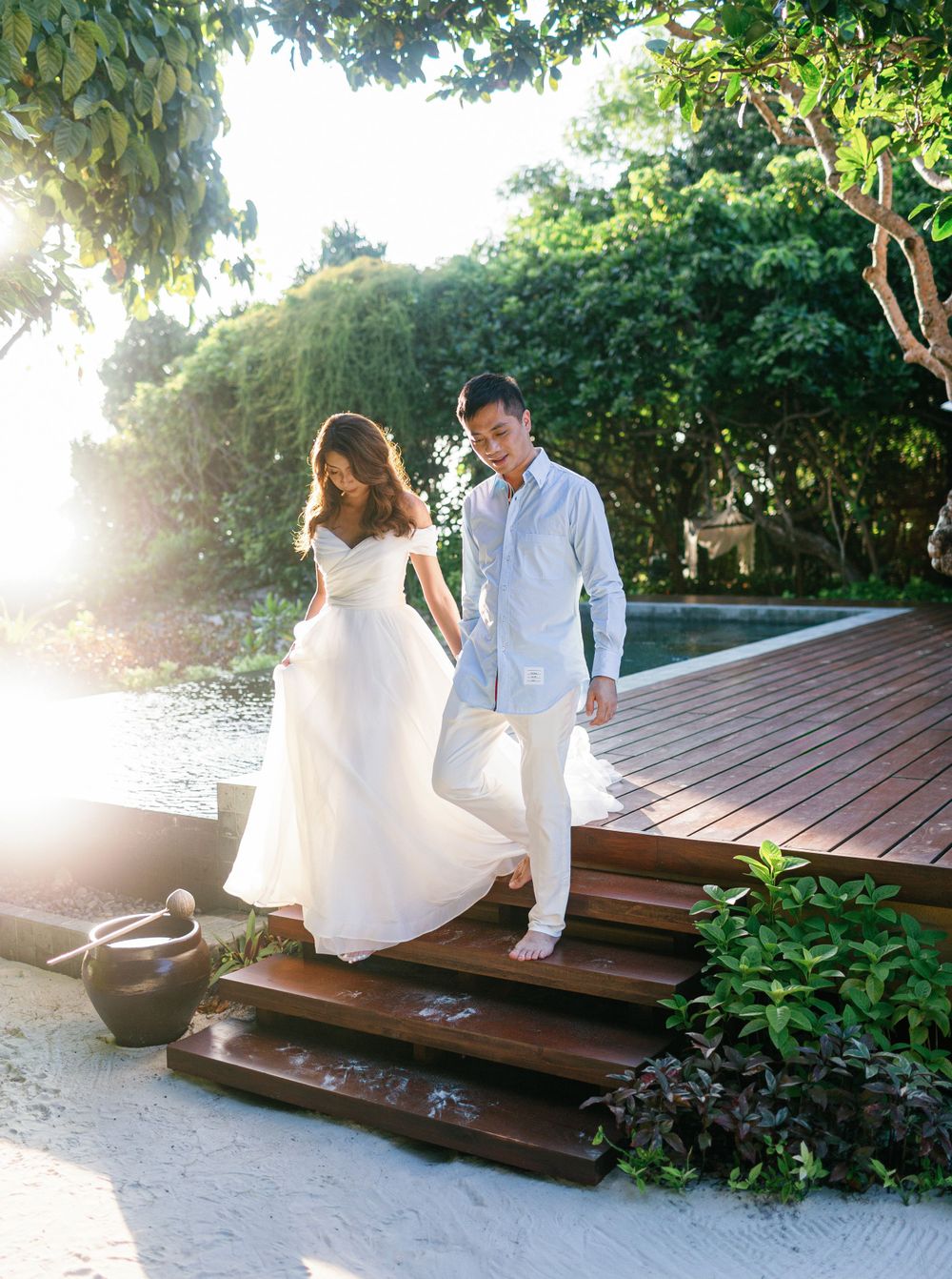 Newlyweds stroll down wooden path by pool at Aman resort in Amanpulo, Philippines.