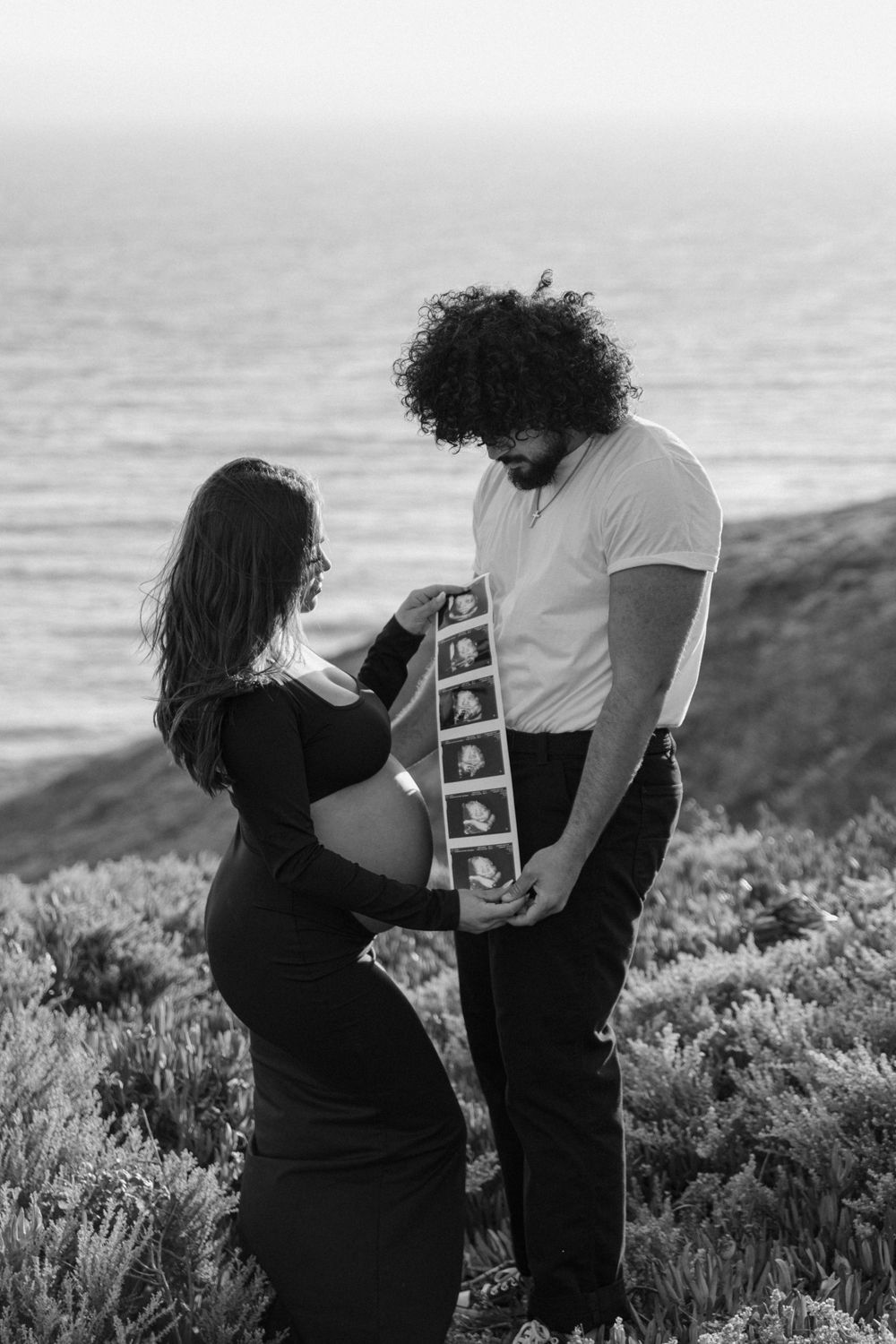 Expectant couple holding ultrasound photos on a scenic cliff overlooking the ocean.