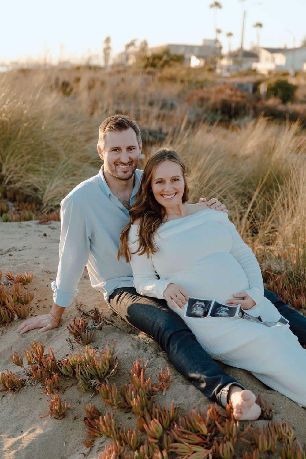 Couple relaxing on coastal dunes during sunset maternity photoshoot.