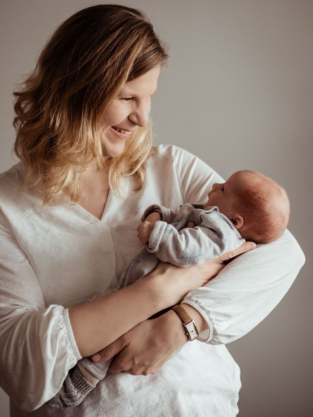 A parent wearing a white top tenderly holds and gazes at their newborn baby wrapped in a gray outfit.