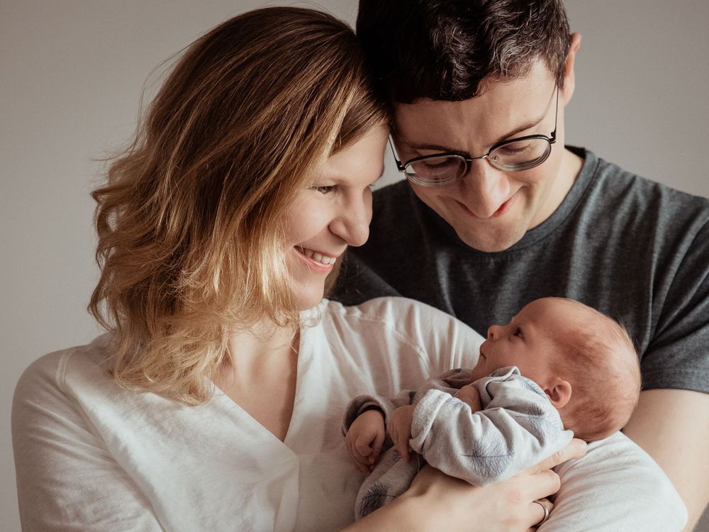 A couple smiling lovingly while holding their newborn baby in a gray and white toned indoor portrait.