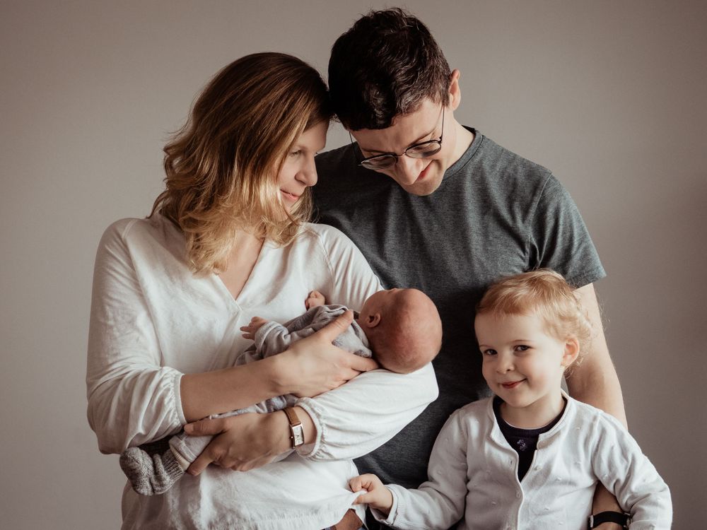 A happy family bonding together in soft sweaters while gathered around their newborn baby on a neutral backdrop.