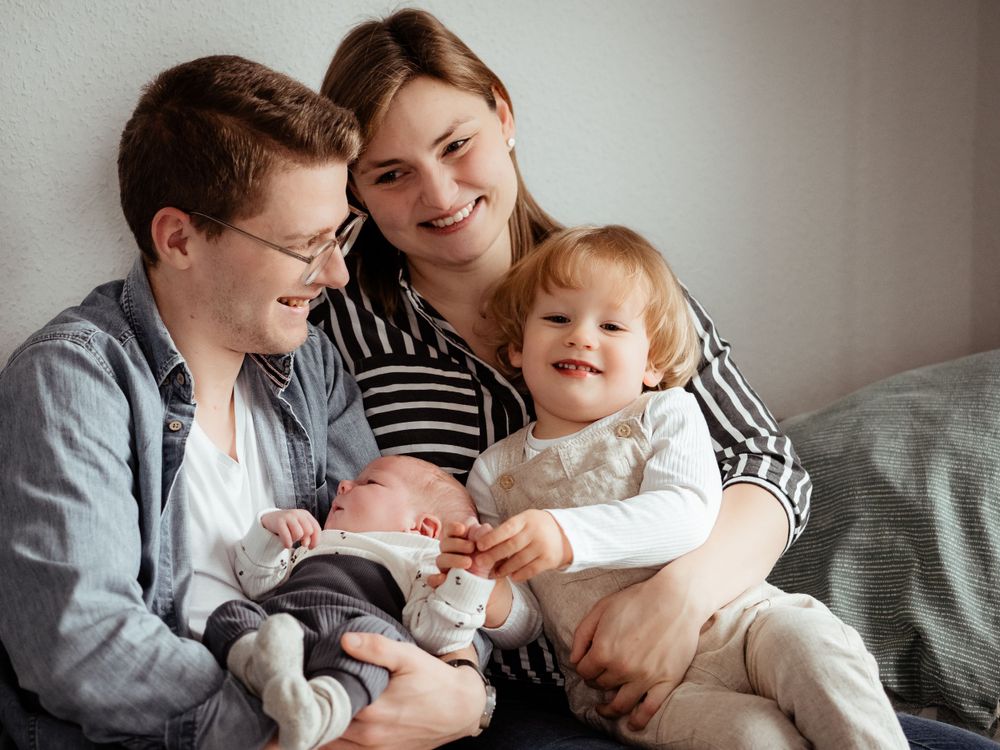A family sits together on a bed smiling while holding a newborn baby and toddler.