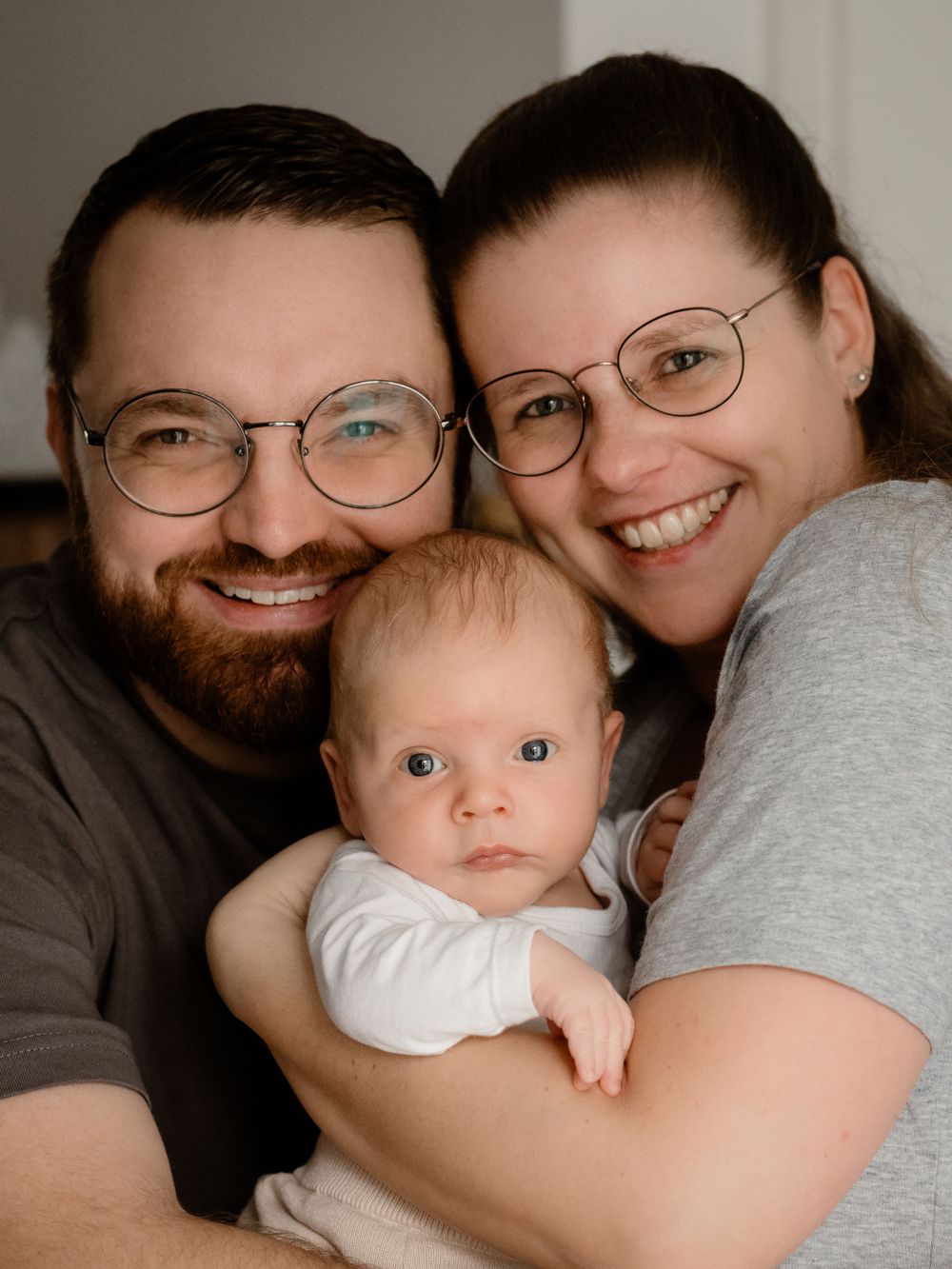 A happy family portrait showing parents wearing glasses embracing their young baby in casual attire.