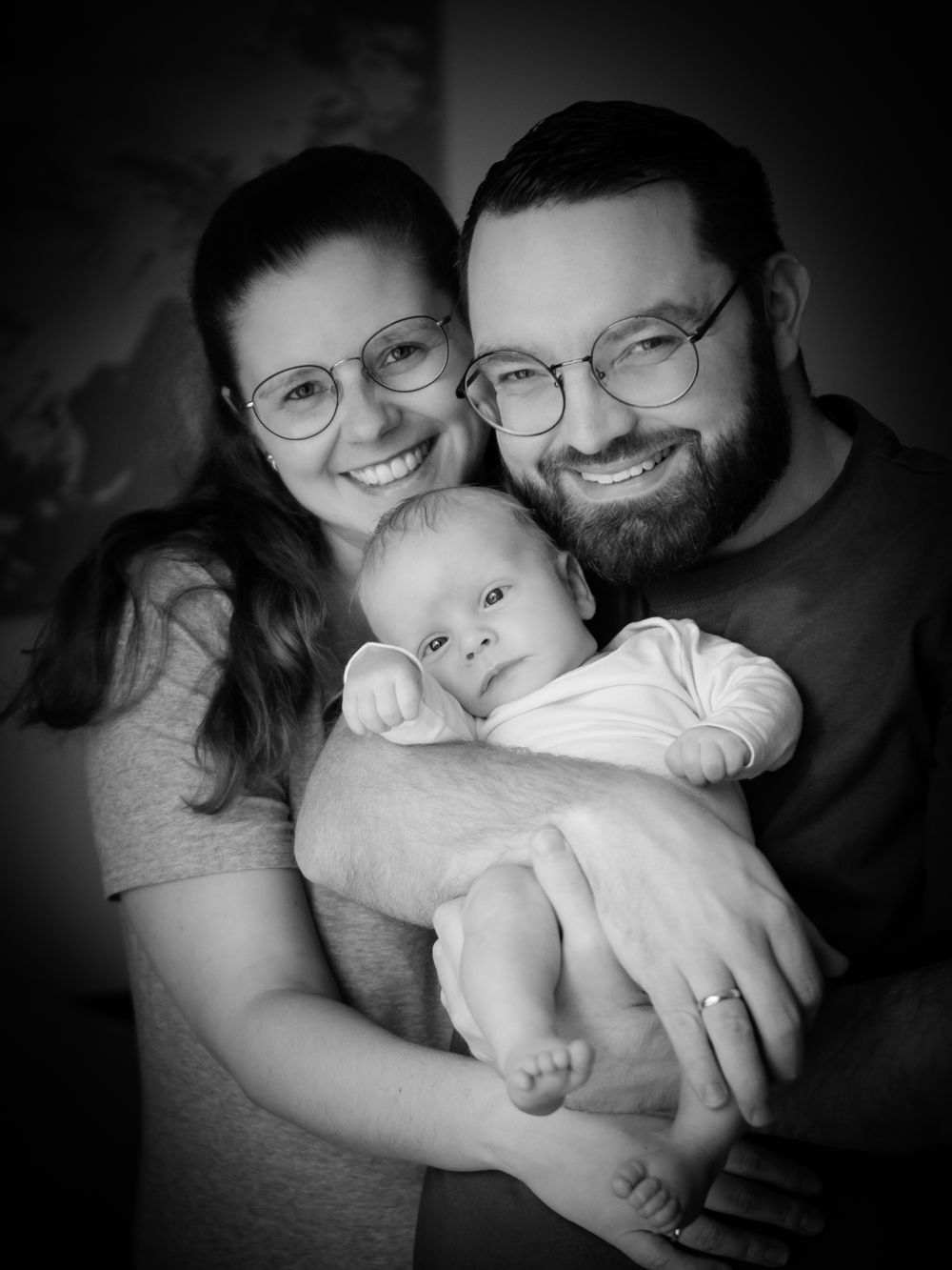 A black and white portrait of a couple in glasses holding their baby.