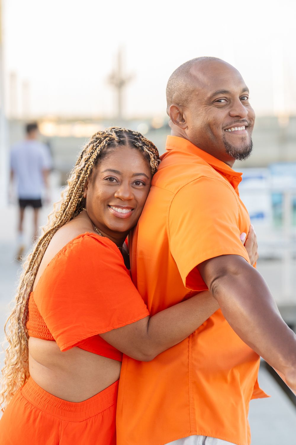 Two people in matching orange outfits share a warm embrace while smiling together outdoors.