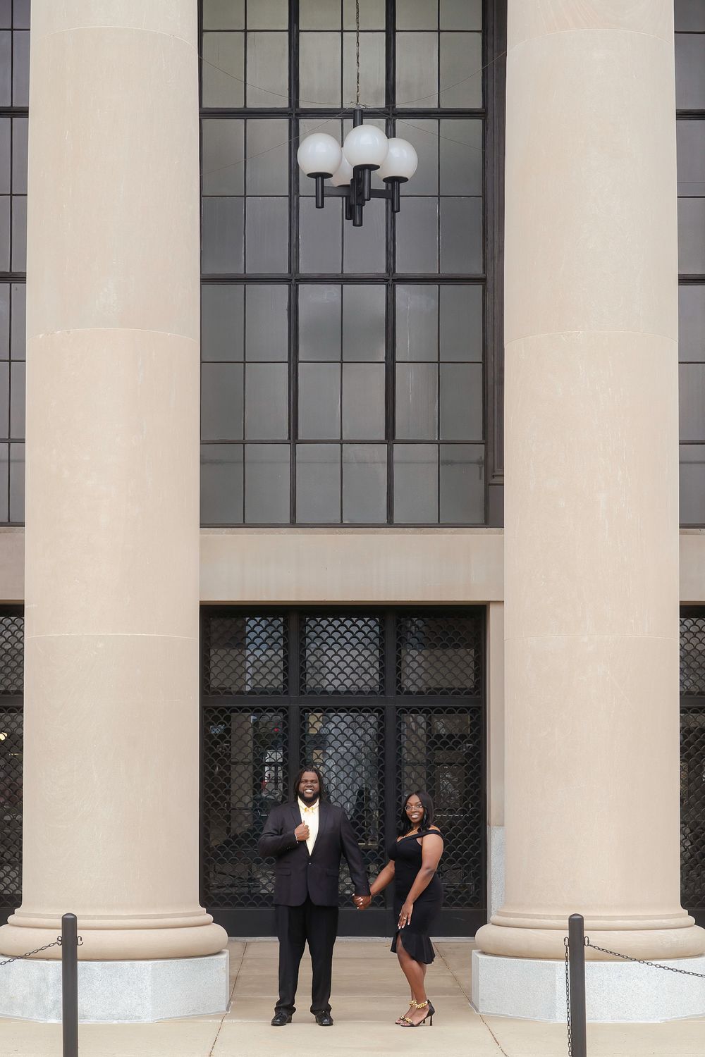 Art deco columns and large windows decorate the entrance of a government building as people stand near the doors.