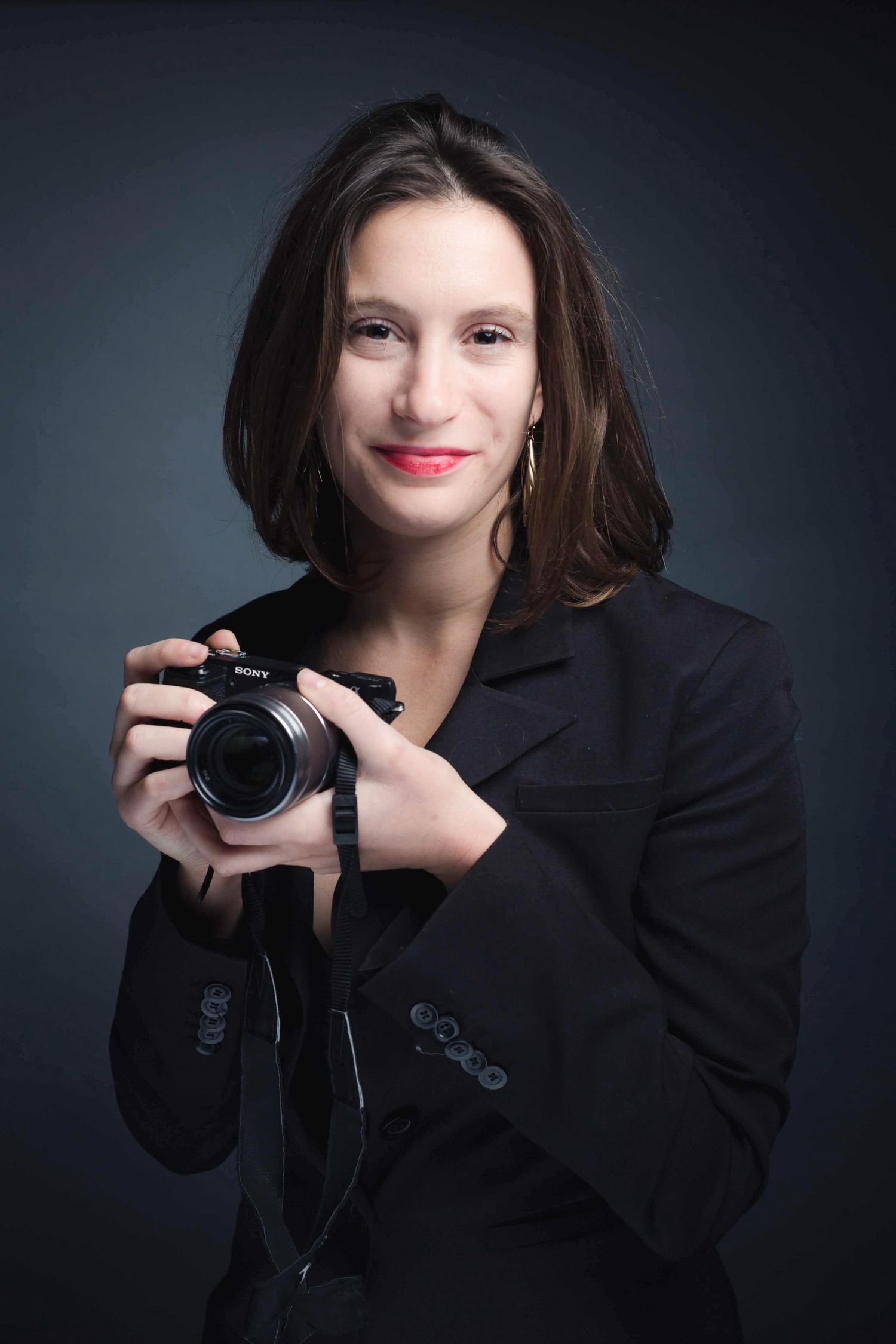 Professional photographer in black blazer holding DSLR camera against dark background.